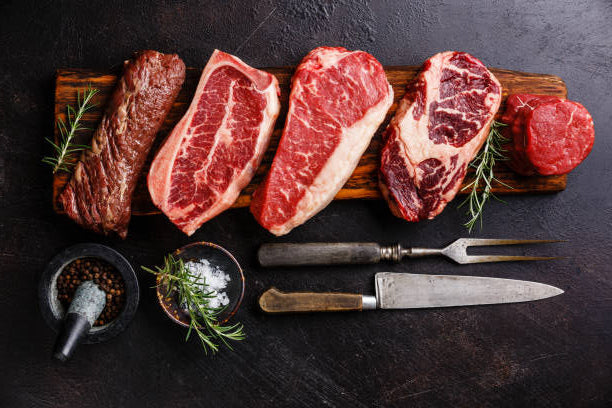 Assorted cuts of raw beef on a wooden board with utensils on a dark surface